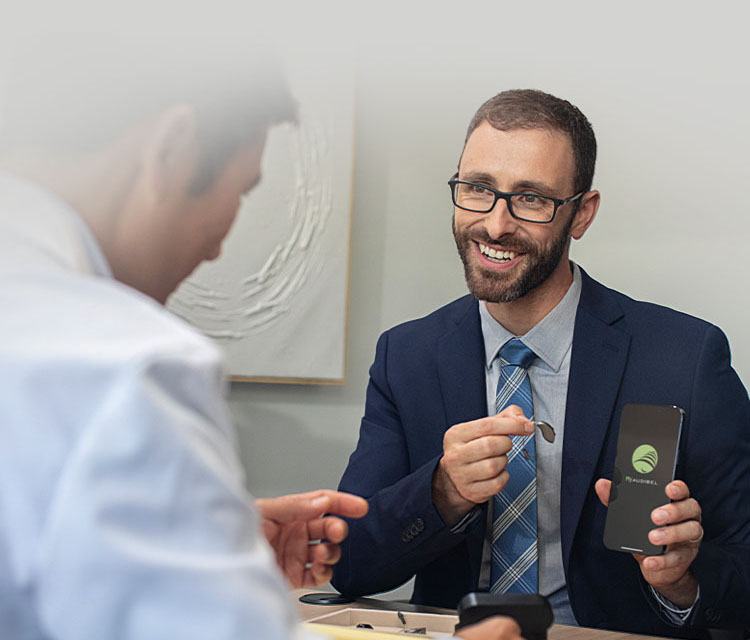 A man holding a hearing aid and a mobile phone with the My Audibel app on it's screen